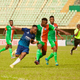 A soccer player for Sporting Lagos in a blue jersey competes for the ball with a Go Round FC player in a green and orange Jersey.